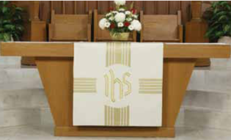 Wooden altar with a white cloth featuring 'IHS' symbol, set against a wooden pew and floral arrangement.